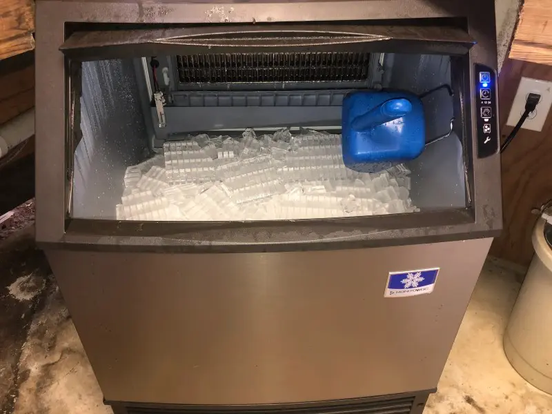A large ice machine with lots of ice and a blue jug.